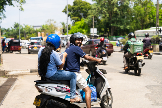 Sri Lanka 19 February 2026 View of a man and woman on a white scooter navigating busy urban traffic with a delivery rider and a red tuk tuk on a sunny day.