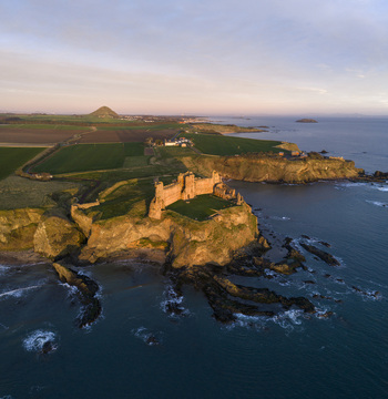 Aerial view of Tantallon Castle on a rugged cliff edge with golden sunset light hitting the stone walls and green fields in North Berwick Scotland United Kingdom.