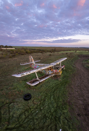 Aerial view of an abandoned biplane covered in colorful graffiti resting in a grassy field under a vibrant sunset sky in Bulgaria. Aerial view of an abandoned biplane covered in colorful graffiti resting in a grassy field under a vibrant sunset sky in Bulgaria.