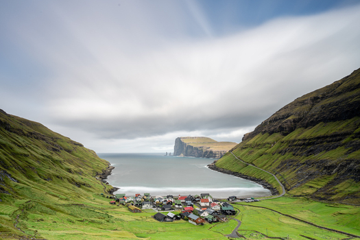 View of the village of Tjornuvik nestled between steep green mountains and a bay with dramatic sea stacks in the Faroe Islands.