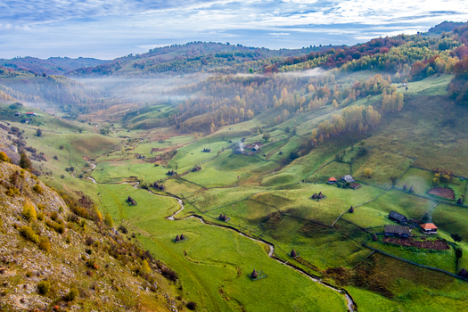 Aerial view of Fundatura Ponorului featuring rolling green hills traditional wooden houses haystacks and a winding stream in the rural landscape of Ohaba Ponor Hunedoara Romania.