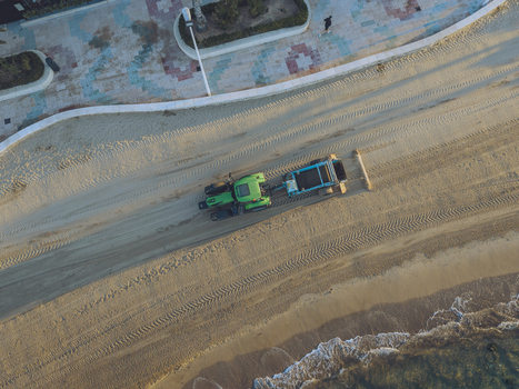 Aerial view of a green tractor raking the sand on a beach next to a paved promenade in Calp Valencian Community Spain. Aerial view of a green tractor raking the sand on a beach next to a paved promenade in Calp Valencian Community Spain.