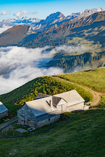 View of a traditional stone mountain hut with a slate roof on a green slope in the Swiss Alps with clouds in the valley and snow capped peaks in the distance in Airolo Ticino Switzerland. View of a traditional stone mountain hut with a slate roof on a green slope in the Swiss Alps with clouds in the valley and snow capped peaks in the distance in Airolo Ticino Switzerland.