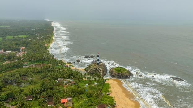 Aerial view of Kapu Light House standing on rocky outcrops along the coastline with lush palm trees and crashing waves in Udupi Karnataka India.