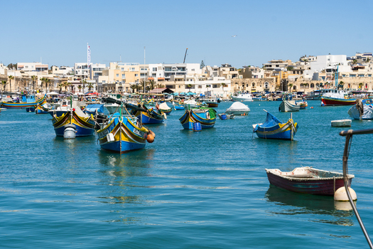 View of traditional colorful Luzzu boats moored in the harbor with coastal architecture and clear blue sky in Marsaskala Southern Region Malta. View of traditional colorful Luzzu boats moored in the harbor with coastal architecture and clear blue sky in Marsaskala Southern Region Malta.
