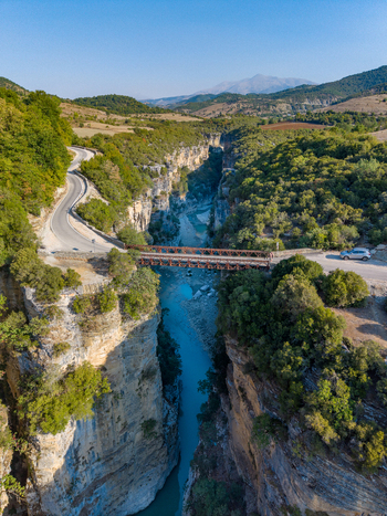 Aerial view of Osumi Canyon featuring a metal bridge crossing the deep gorge a winding road lush green vegetation and turquoise river water in Dhores Berat County Albania.