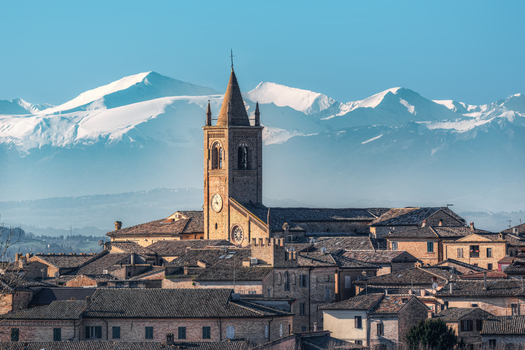 View of the historic Collegiate Church of Santa Maria Assunta rising above stone rooftops with snow capped mountains in the background. Montecassiano Marche Italy. View of the historic Collegiate Church of Santa Maria Assunta rising above stone rooftops with snow capped mountains in the background. Montecassiano Marche Italy.