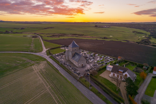 Aerial view of a historic stone church and its surrounding cemetery set amidst rural fields under a vibrant sunset sky in Audresselles Hauts de France France.