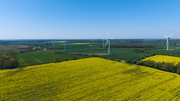 Aerial view of the New Albion Wind Farm with white turbines standing amidst vibrant yellow rapeseed fields and green pastures under a clear blue sky in Rushton England United Kingdom. Aerial view of the New Albion Wind Farm with white turbines standing amidst vibrant yellow rapeseed fields and green pastures under a clear blue sky in Rushton England United Kingdom.