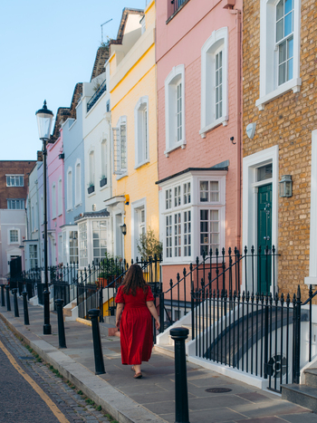 View of a woman in a red dress walks along a sidewalk lined with pastel colored terraced houses under a clear sky creating a vibrant street scene London United Kingdom. View of a woman in a red dress walks along a sidewalk lined with pastel colored terraced houses under a clear sky creating a vibrant street scene London United Kingdom.