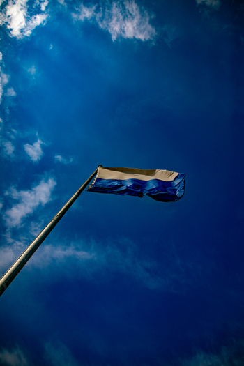 View of a blue and white flag waving on a tall flagpole against a vibrant blue sky with scattered white clouds in Offenburg Baden Wurttemberg Germany. View of a blue and white flag waving on a tall flagpole against a vibrant blue sky with scattered white clouds in Offenburg Baden Wurttemberg Germany.