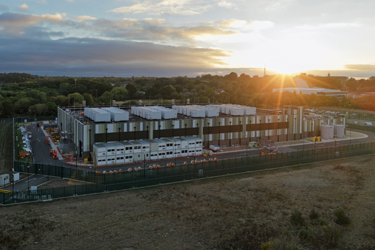 Aerial view of a modern data center facility featuring rooftop cooling units and a fenced perimeter during golden hour in the United Kingdom. Aerial view of a modern data center facility featuring rooftop cooling units and a fenced perimeter during golden hour in the United Kingdom.