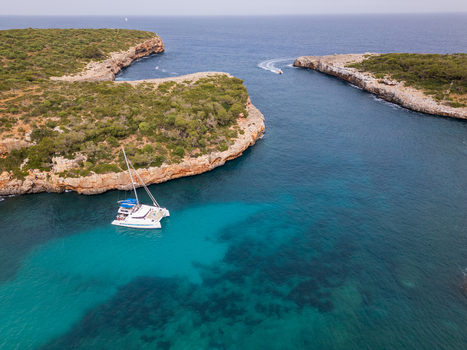 Aerial view of Cala Sa Nau with a white catamaran anchored in turquoise water and people on deck near rocky cliffs and green vegetation in Felanitx Balearic Islands Spain. Aerial view of Cala Sa Nau with a white catamaran anchored in turquoise water and people on deck near rocky cliffs and green vegetation in Felanitx Balearic Islands Spain.
