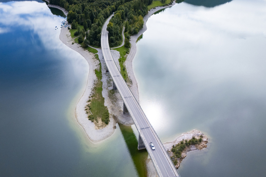 Aerial view of a bridge cutting through the tranquil lake mirroring the sky connecting lush green landmasses Sylvensteinspeicher Bayern Germany. Aerial view of a bridge cutting through the tranquil lake mirroring the sky connecting lush green landmasses Sylvensteinspeicher Bayern Germany.
