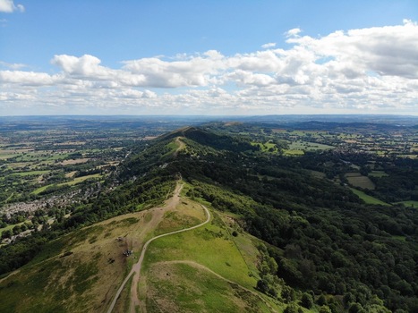 Aerial view of the Malvern Hills featuring rolling green ridges winding footpaths and lush woodland under a bright blue sky with scattered clouds in Malvern England United Kingdom.