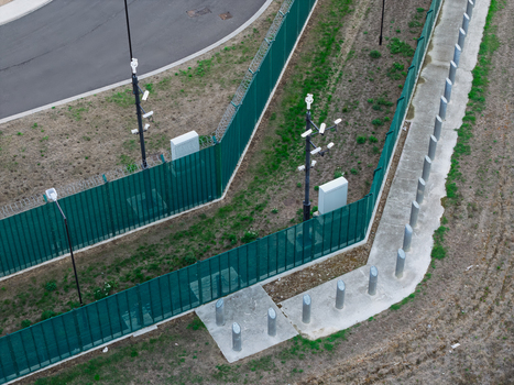 Aerial view of a data center perimeter featuring green security fencing multiple surveillance cameras mounted on poles and concrete bollards along a paved walkway in the United Kingdom. Aerial view of a data center perimeter featuring green security fencing multiple surveillance cameras mounted on poles and concrete bollards along a paved walkway in the United Kingdom.