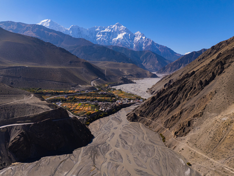Aerial view of a braided river winding through a deep Himalayan valley with snow capped peaks terraced fields and a small village in Nepal.