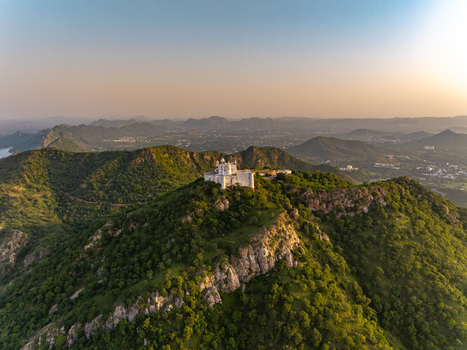 Aerial view of Sajjangarh Monsoon Palace perched on a lush green hilltop surrounded by the Aravalli mountain range during golden hour in Udaipur Rajasthan India.