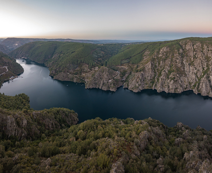 Aerial view of the Ribeira Sacra canyon with the Sil River winding through steep cliffs covered in lush green forests under a soft twilight sky in Lugo Orense Spain. Aerial view of the Ribeira Sacra canyon with the Sil River winding through steep cliffs covered in lush green forests under a soft twilight sky in Lugo Orense Spain.