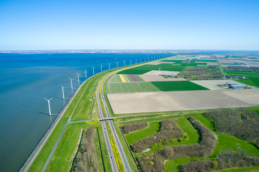 Aerial view of wind turbines lining the dike along the coast with agricultural fields and a highway in the Netherlands.