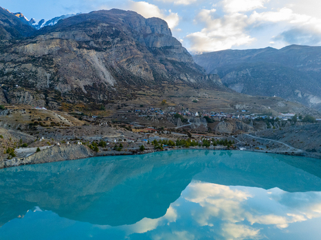 Aerial view of a turquoise glacial lake reflecting the sky surrounded by rugged Himalayan mountains and a small village in Nepal.