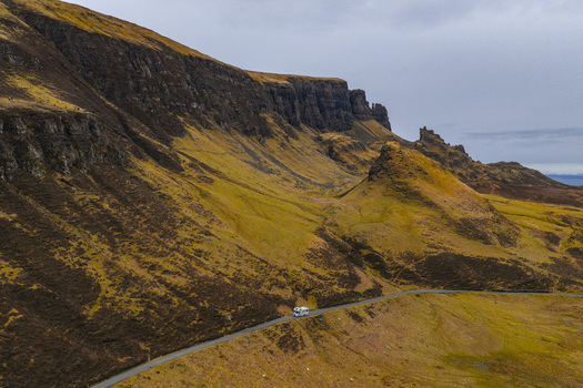 Aerial view of The Quiraing with a white campervan driving on a winding road through the dramatic rocky landscape and green slopes Isle of Skye Scotland United Kingdom. Aerial view of The Quiraing with a white campervan driving on a winding road through the dramatic rocky landscape and green slopes Isle of Skye Scotland United Kingdom.