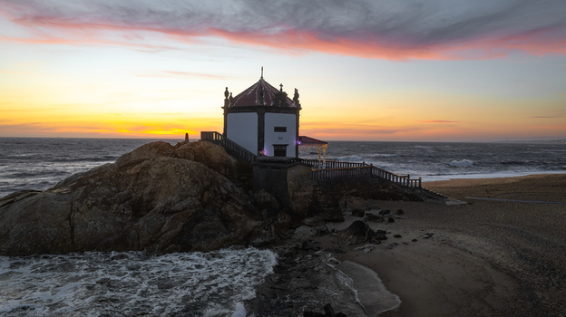 Aerial view of Capela do Senhor da Pedra on a rocky outcrop at the beach during a vibrant sunset with crashing waves in Miramar Porto Portugal.