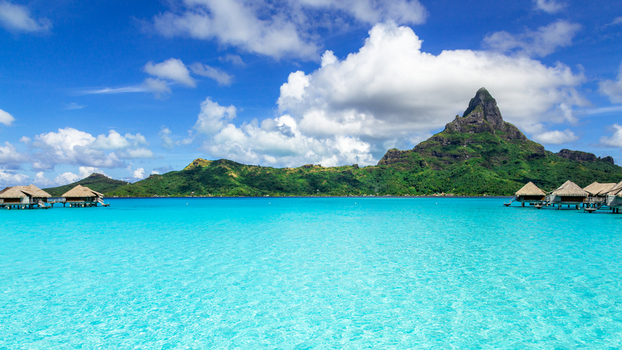 View of overwater bungalows in a turquoise lagoon with Mount Otemanu in the background under a blue sky in Bora Bora Leeward Islands French Polynesia.