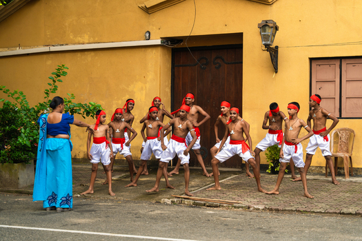 Sri Lanka 04 February 2026 View of young boys in traditional attire practicing a folk dance with a female instructor in front of a historic yellow building.