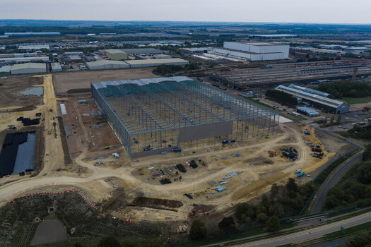 Aerial view of a large industrial construction site featuring the steel framework of a warehouse under development with surrounding dirt roads and heavy machinery in the United Kingdom. Aerial view of a large industrial construction site featuring the steel framework of a warehouse under development with surrounding dirt roads and heavy machinery in the United Kingdom.