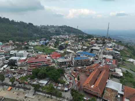 Aerial view of the town near Sarangan Lake with dense buildings red tiled roofs and lush green hills under a cloudy sky in Plaosan East Java Indonesia. Aerial view of the town near Sarangan Lake with dense buildings red tiled roofs and lush green hills under a cloudy sky in Plaosan East Java Indonesia.