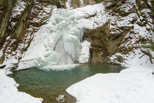 Aerial view of the frozen Casoca Waterfall cascading into a clear pool surrounded by snow covered rocks and forest in winter in Casoca Buzau County Romania.
