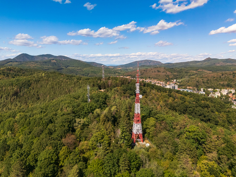 Aerial view of a red and white broadcast tower rising above forested hills and the cityscape of Salgotarjan Hungary under a blue summer sky with scattered clouds. Aerial view of a red and white broadcast tower rising above forested hills and the cityscape of Salgotarjan Hungary under a blue summer sky with scattered clouds.