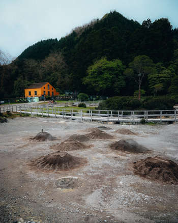 View of steaming fumaroles bubble from the earth in a geothermal field contrasting with the vibrant orange building against the backdrop of lush green hills Furnas Azores Portugal.