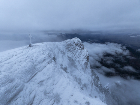 Aerial view of the snowy Pyrenees mountain peak featuring a prominent cross at the summit surrounded by thick clouds in Spain. Aerial view of the snowy Pyrenees mountain peak featuring a prominent cross at the summit surrounded by thick clouds in Spain.