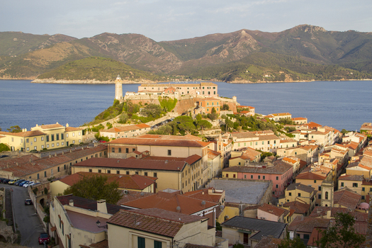 View of sun kissed buildings with red tiled roofs cascade down the hillside towards the shimmering sea crowned by a historic fortress Portoferraio Elba Tuscany Italy. View of sun kissed buildings with red tiled roofs cascade down the hillside towards the shimmering sea crowned by a historic fortress Portoferraio Elba Tuscany Italy.