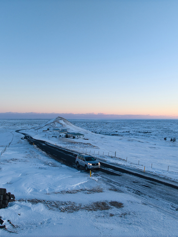 Aerial view of a silver SUV driving on a snow covered road through a vast icy landscape under a clear pale sky at sunset in Kirkjubaejarklaustur Skaftarhreppur Iceland. Aerial view of a silver SUV driving on a snow covered road through a vast icy landscape under a clear pale sky at sunset in Kirkjubaejarklaustur Skaftarhreppur Iceland.