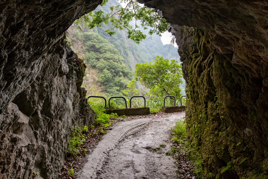 View of a narrow unpaved trail carved through mossy rock walls with a metal guardrail and lush green vegetation in Taroko National Park Xiulin Township Hualien County Taiwan.
