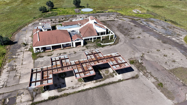 Aerial view of an abandoned building with a red tiled roof and a rusted metal canopy structure surrounded by cracked pavement and green fields in iNtabazwe Free State South Africa. Aerial view of an abandoned building with a red tiled roof and a rusted metal canopy structure surrounded by cracked pavement and green fields in iNtabazwe Free State South Africa.