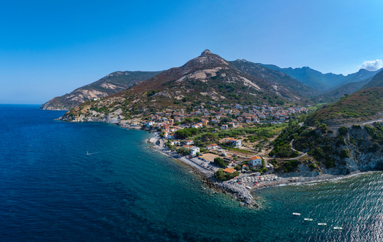 Aerial view of Pomonte village nestled between the rugged mountains and the blue Tyrrhenian Sea featuring stone houses coastal roads and rocky beaches in Pomonte Tuscany Italy.