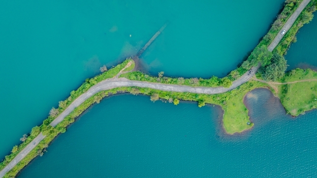 Aerial view of Upper Vaitarana Dam with a winding road cutting through the vibrant turquoise reservoir surrounded by lush green landscape in Ondli Maharashtra India.