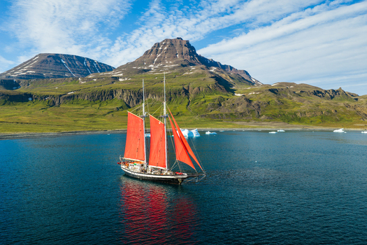 Aerial view of a sailboat with striking red sails navigating through a calm bay surrounded by rugged mountains and scattered icebergs in Greenland. Aerial view of a sailboat with striking red sails navigating through a calm bay surrounded by rugged mountains and scattered icebergs in Greenland.