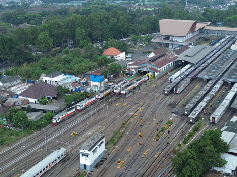 Aerial view of Stasiun Malang Kota Baru with multiple passenger trains parked on tracks surrounded by lush trees and urban buildings in Kecamatan Klojen Jawa Timur Indonesia.