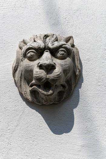 View of a stone lion head sculpture with an open mouth mounted on a white textured wall under bright sunlight Monreal Rhineland Palatinate Germany. View of a stone lion head sculpture with an open mouth mounted on a white textured wall under bright sunlight Monreal Rhineland Palatinate Germany.