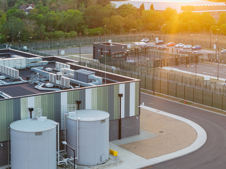 Aerial view of a modern data center facility with large industrial water tanks and HVAC units on the roof surrounded by security fencing and a parking area at sunset in the United Kingdom. Aerial view of a modern data center facility with large industrial water tanks and HVAC units on the roof surrounded by security fencing and a parking area at sunset in the United Kingdom.