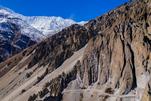 Aerial view of rugged rocky cliffs and a winding mountain trail leading towards snow capped Himalayan peaks under a clear blue sky in Nepal.