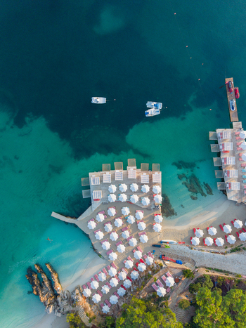 Aerial view of the Ksamil beach resort featuring rows of white umbrellas on wooden decks turquoise water and small boats moored nearby in Ksamil Vlore County Albania. Aerial view of the Ksamil beach resort featuring rows of white umbrellas on wooden decks turquoise water and small boats moored nearby in Ksamil Vlore County Albania.