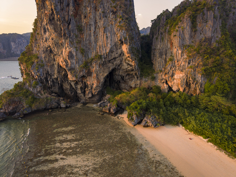 Aerial view of rugged cliffs touched by golden sunlight meet the serene beach nestled beside lush greenery Krabi Thailand.