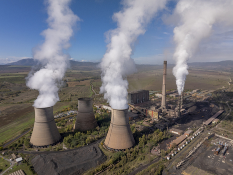 Aerial view of a coal fired power plant with cooling towers and tall chimneys emitting steam into the clear sky over the rural landscape of Bulgaria.