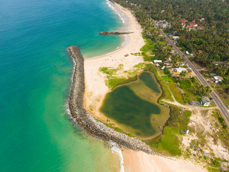 Aerial view of the sandy coastline with a stone breakwater turquoise ocean waves and lush palm trees along the coastal road Peraliya Southern Province Sri Lanka. Aerial view of the sandy coastline with a stone breakwater turquoise ocean waves and lush palm trees along the coastal road Peraliya Southern Province Sri Lanka.